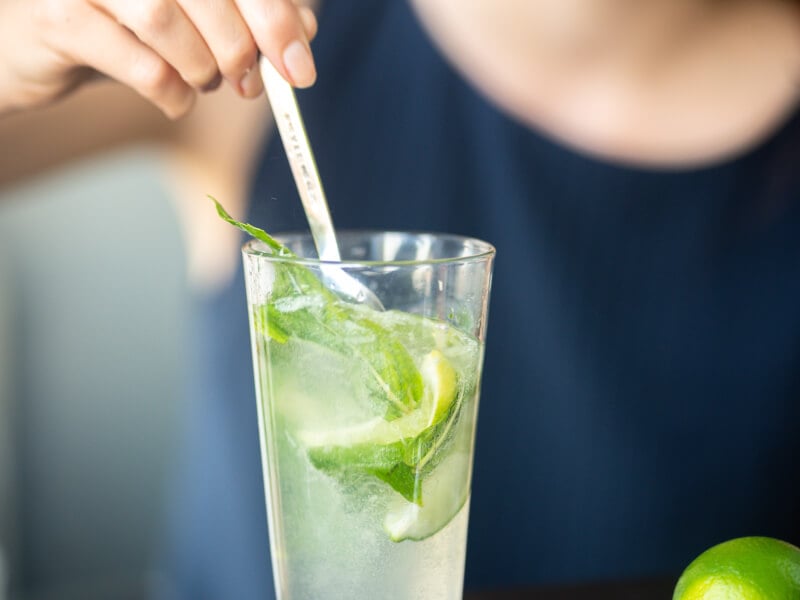 A hand stirs a spoon into a mojito cocktail containing lime and fresh mint